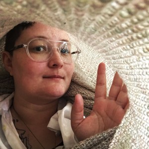 Portrait of D. Allen, a white queer person with glasses and short hair, looking at the camera from inside a handknit textile tube. 